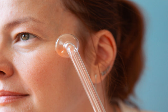 woman using darsonval electric device on cheek gazing away in bright studio closeup, concept of skincare routine, cosmetology, aesthetic medicine