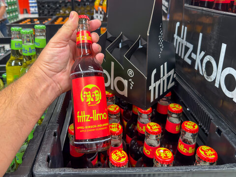 Berlin, Germany July 29, 2026. A hand holds a red labeled fritz limo soda bottle flavored with apple cherry and elderberry at a store. Crates of fritz kola and fritz limo bottles fill the blurred back
