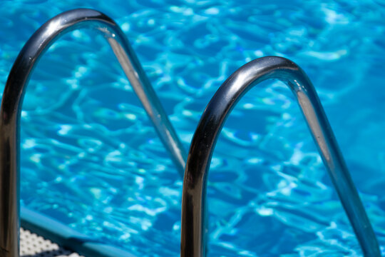 stainless steel swimming pool ladder entering blue water with ripples, deck and sun loungers in background closeup, concept of hotel resort, vacation, poolside relaxation