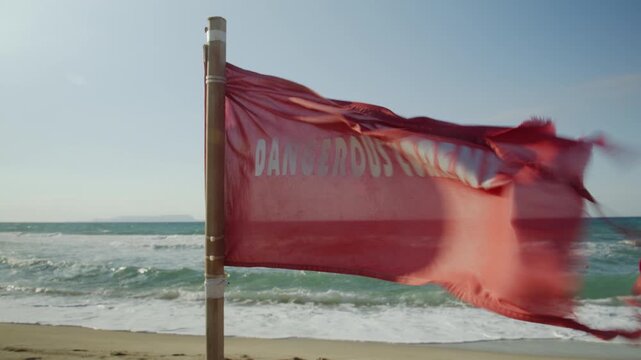 Close-up of a red warning flag for dangerous currents flapping in the strong wind on a beach with rough ocean waves.