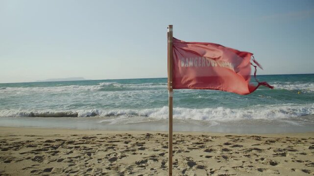 A red warning flag flutters in the wind on an empty sandy beach, alerting swimmers to dangerous ocean currents and waves.