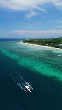 Vertical drone follows a longboat, ascends, and pulls back over deep blue water and reef flats near Dumaluan Beach in Panglao, Bohol