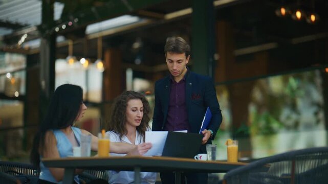 Three business professionals are engaged in an outdoor meeting at a cafe. They review documents and collaborate on a laptop in an open-air setting.