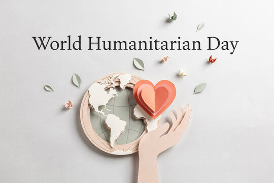 Clean studio flat lay concept for world humanitarian day showing paper cutout human hand holding red heart next to minimalist earth globe disk and small origami paper cranes on gray backdrop.
