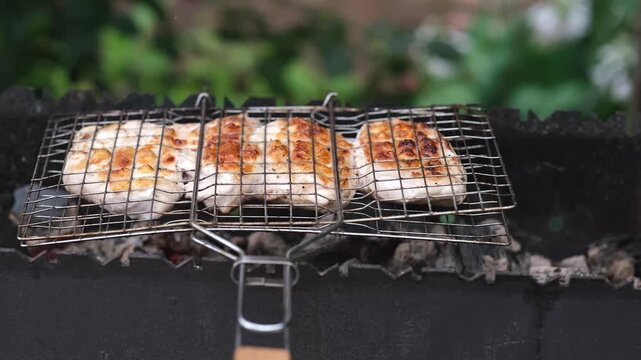 Grilled meat cooking in a barbecue grill basket on a backyard barbecue with green bushes in the background. Summer outdoor cooking.