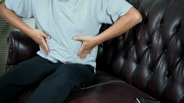 Stomach ache, stomach cramps, heartburn, digestive problems. A young Asian man is holding his stomach and grimacing in pain while sitting on the couch at home.
