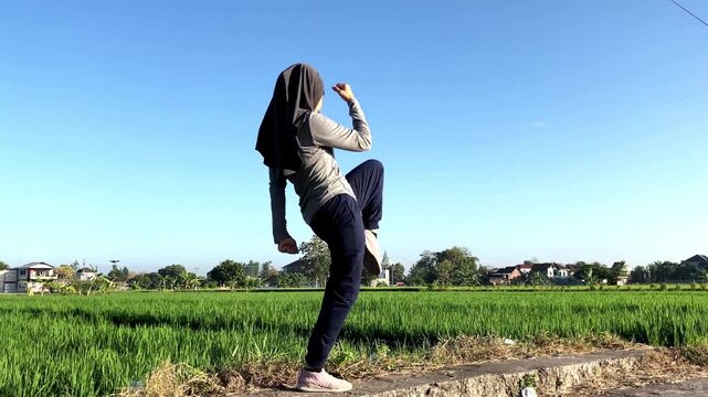 An active Muslim woman wearing a hijab practices shadow boxing and martial arts moves in a beautiful green rice field under a clear blue sky. Ideal for fitness, self-defense, and wellness concepts.