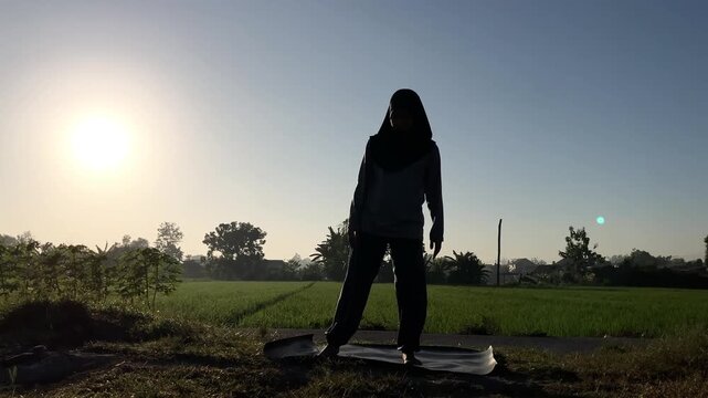 Slow motion silhouette of a Muslim woman in a hijab doing the triangle yoga pose (Trikonasana) on a mat outdoors. Beautiful golden sunrise over a lush green rice field. Perfect for wellness themes.