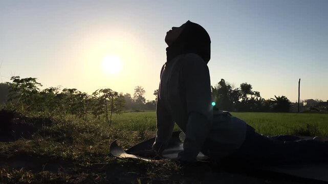 Slow motion clip of a Muslim woman in a hijab doing cobra yoga pose on a mat outdoors. Beautiful golden sunrise backlight over a green field. Perfect for wellness and fitness concepts.