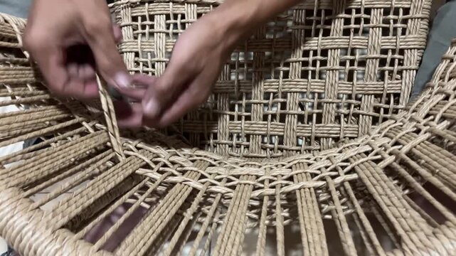 Slow motion close-up shot of artisan hands weaving natural fiber cord to create a traditional chair seat. Detailed view of the craftsmanship and handmade furniture making process.