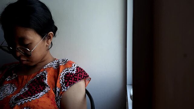 Cinematic clip of a thoughtful Asian woman in glasses looking down near a window. Dark, moody natural lighting creates an introspective atmosphere. Ideal for emotional or everyday lifestyle themes.