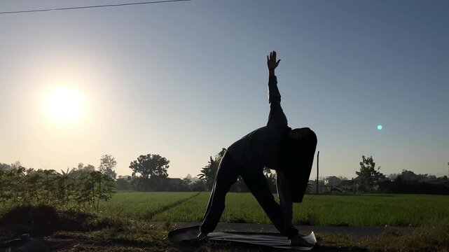Slow motion silhouette of a Muslim woman in a hijab doing the triangle yoga pose (Trikonasana) on a mat outdoors. Beautiful golden sunrise over a lush green rice field. Perfect for wellness themes.