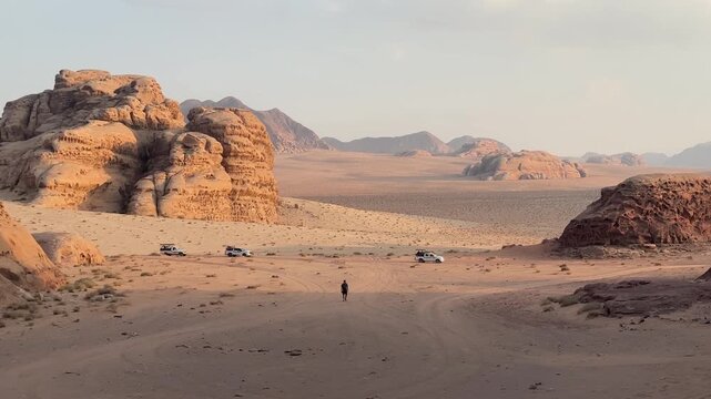 Cinematic footage of a man walking through the dramatic red desert landscape of Wadi Rum with three trucks following behind, capturing the raw adventure and epic scale of one of Jordan's most iconic d