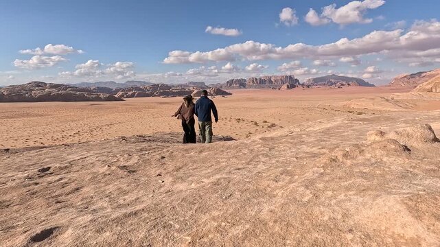 Breathtaking footage of a couple walking along a dramatic cliff edge high above the vast red desert landscape of Wadi Rum, soaking up the extraordinary otherworldly scenery of one of Jordan's most ico