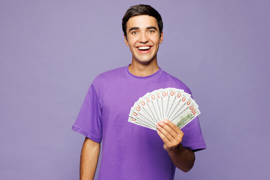 Young happy Caucasian man he wear violet t-shirt casual clothes hold in hand fan of cash money in dollar banknotes isolated on plain pastel light purple background studio portrait. Lifestyle concept.