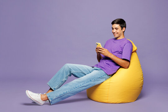Full body young happy man he wear violet t-shirt casual clothes sit in bag chair hold in hand use mobile cell phone isolated on plain pastel light purple background studio portrait. Lifestyle concept.
