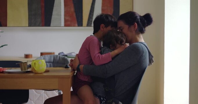 Amused mother balances attention between a clingy baby and older sibling at the dining table at home, easing playful rivalry with patience, affection and family harmony