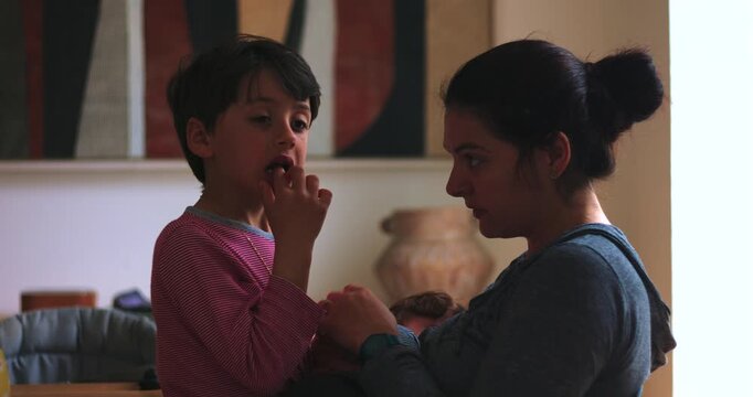 Young boy reveals a wobbly tooth while his mother examines his smile at home with a baby in her arms, celebrating healthy development, growing up and parental care