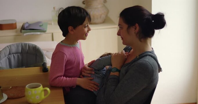 Breastfeeding mother talks with older son while nursing toddler at the breakfast table at home, capturing candid family interaction, maternal care and sibling closeness
