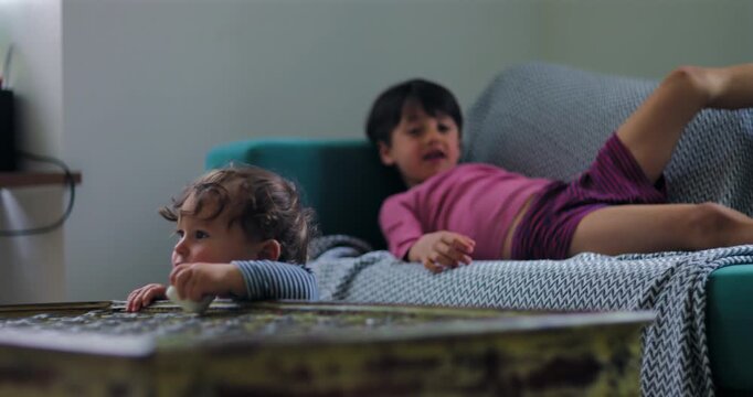 Toddler reaches from coffee table toward sofa and attempts first steps at home as older brother claps encouragement, celebrating mobility, confidence and a childhood milestone