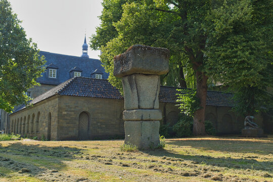 A distinctive stone monument stands in a sunlit grassy area. Historic stone buildings and green trees frame the background, depicting a quiet European scene - Soest, Westphalia, Germany