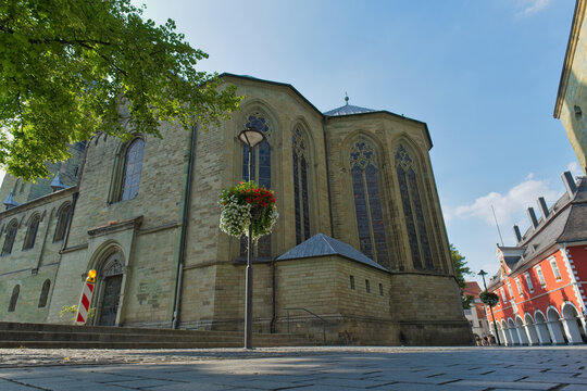 A large, historic stone church with arched windows stands on a sunny day. A decorative flower basket and paved square are in the foreground - Soest, Westphalia, Germany