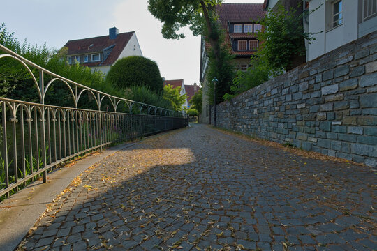 A sunlit, narrow cobblestone path winds uphill through a European town, flanked by a metal railing and stone wall - Soest, Westphalia, Germany