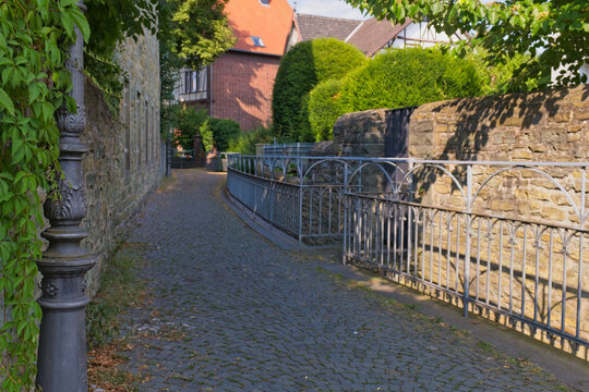 A scenic cobblestone alley ascends between stone walls, featuring an ornate lamppost, green foliage, and distant historic buildings - Soest, Westphalia, Germany
