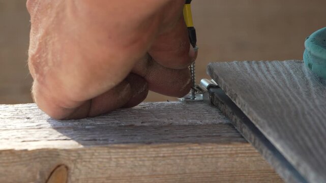 Construction worker secures metal decking clip into wooden joist using a drill, building a patio. Close-up static shot.