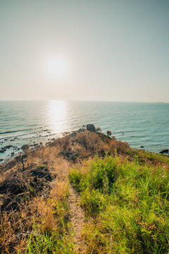 A narrow dirt path leading towards a rocky headland overlooking the gleaming sea at sunset