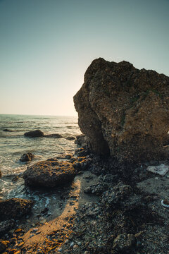 A massive textured boulder standing on a wet coastal beach during golden hour with gentle sea waves.