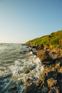 Gentle sea waves washing against a rocky coastal shore with a lush green grassy hill rising on the right.