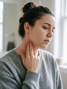 Young woman with swollen neck and sore throat, woman touching a red swollen area on her neck in bright home interior, close up portrait, natural daylight, realistic healthcare photography, sore throat