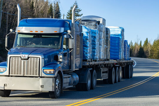 Quebec, Canada, 24 April 2026 : A blue Western Star semi-truck with a flatbed trailer transports wrapped cargo along a paved road.