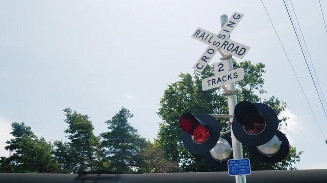 Railway crossing signal lights flashing while freight train passes on tracks in sunny outdoor setting.