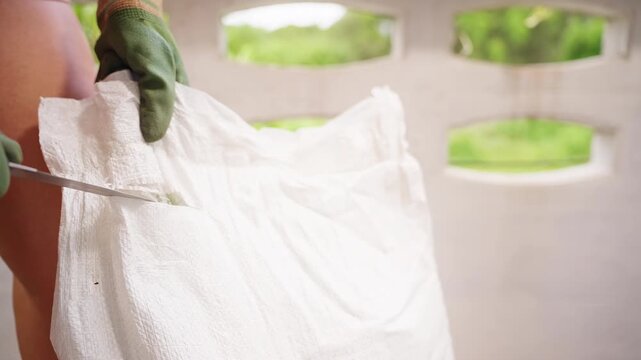 Woman Wearing Protective Gloves Uses a Knife to Cut across the Top of a White Soil Bag, Opening Gardening Supplies for Plant Cultivation