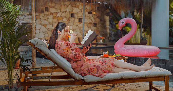 Indian young adult relax woman sitting on sun lounger chair read new novel book enjoy picnic trip day happy girl hold glass drink casual juice do self care spend time have fun joy pool edge place