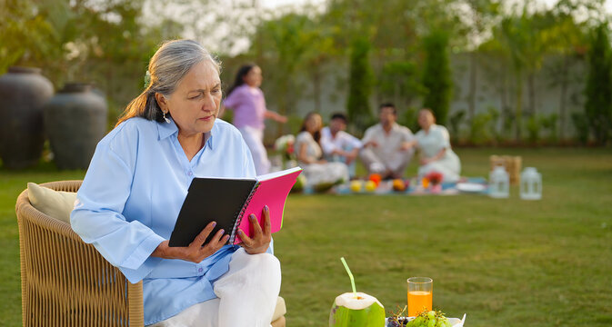 Indian old aged relax woman mom sitting on chair hold book read story novel outdoor house place happy male female man group talk speak spend quality time enjoy new trip day open in air garden lawn