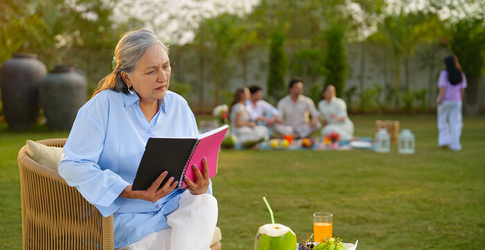 Indian old aged relax woman mom sitting on chair hold book read story novel outdoor house place happy male female man group talk speak spend quality time enjoy new trip day open in air garden lawn