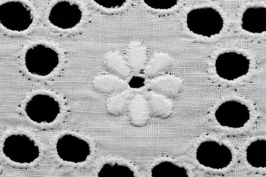 Extreme close-up macro shot of a single embroidered daisy flower surrounded by round cutwork eyelets on white cotton broderie anglaise, isolated on black background.
