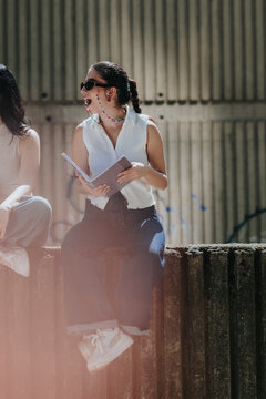 A student in sunglasses laughs while holding an open notebook as she sits on a concrete wall. Another student sits beside her and they chat on a sunny urban ledge.