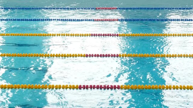 Close-up of blue pool water with bright sun glare and ripples. Yellow, blue, and red floating lane dividers stretch across the frame. Fresh and dynamic swimming pool atmosphere 4k video
