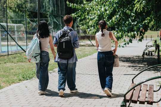 Three students walk along a paved campus path carrying backpacks. They stroll side by side past a sports field and shaded benches on a sunny day.
