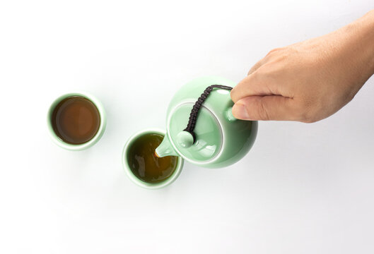 Hand pouring hot tea from green ceramic celadon teapot into teacup isolated on white background
