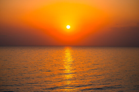 Wide angle shot of bright glowing sun setting over sea horizon casting golden light on calm water.