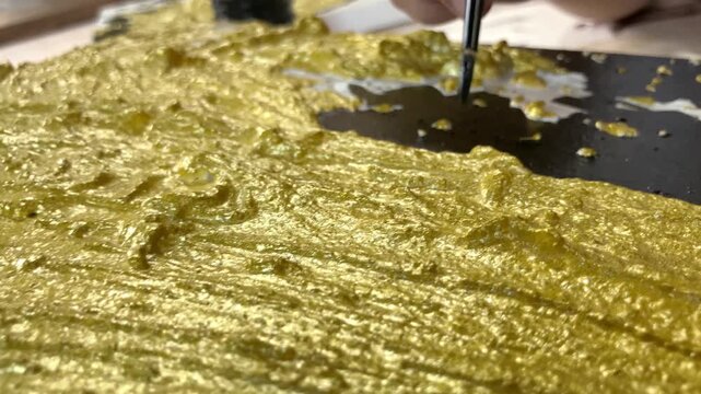 Slow motion close up of a female artist wearing glasses meticulously applying gold leaf onto a black textured canvas with a brush. Creating modern abstract art in a studio workspace.
