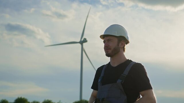 Man in hard hat and overalls looks through binoculars at wind turbines during sunset. Renewable energy, clean electricity, wind power inspection, eco-friendly technology, sustainable industry