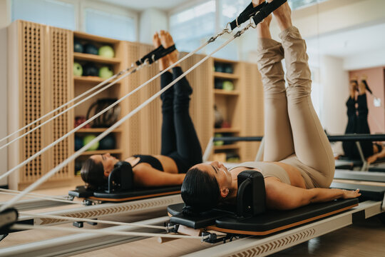 Two women perform a pilates reformer workout on reformer machines with legs extended upward. The pair practice controlled leg exercises in a bright, modern fitness studio environment.