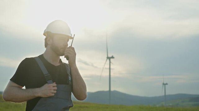 Man in hard hat and overalls speaks into radio near wind turbines on sunny day. Renewable energy, clean electricity, wind power inspection, eco-friendly technology, sustainable industry, teamwork,