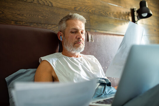 Middle aged man reviewing bills on laptop in bed at home